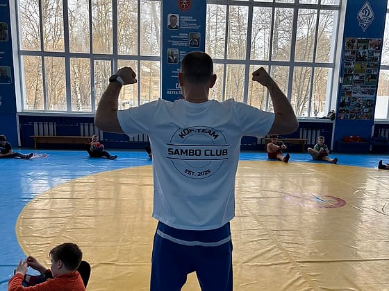 Тренер в белой футболке Kop.team Sambo Club в зале борьбы, вид со спины